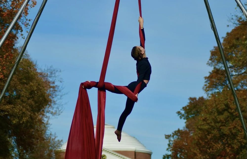 An aerial dance performer, dressed in all black, performs a dance in the air with a red aerial silk on UVA's South Lawn.