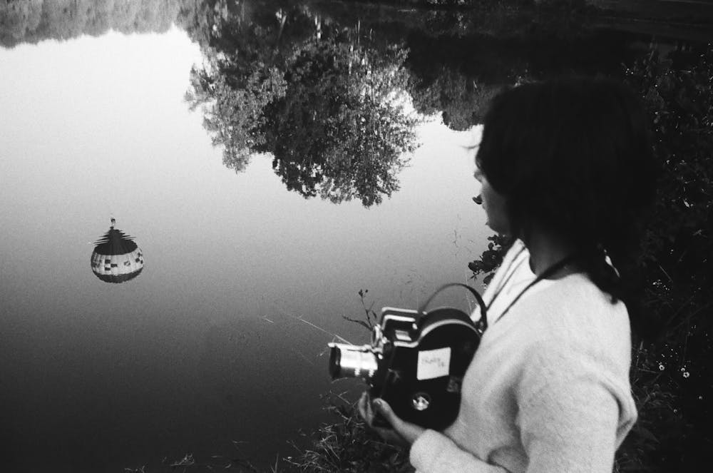 A black-and-white photo of Quintero holding a camera and looking out onto the reflection of a hot air balloon in a pond.